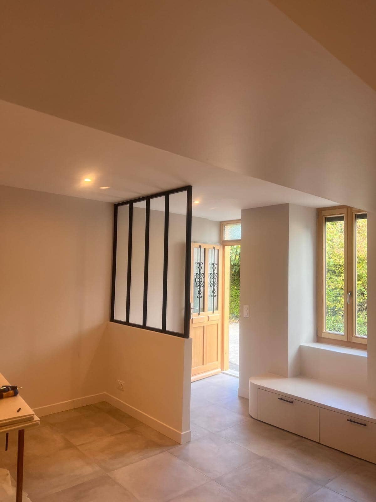Entrée d'une maison rénovée : verrière d'atelier à montants noirs, porte en bois vitrée, banc sous la fenêtre